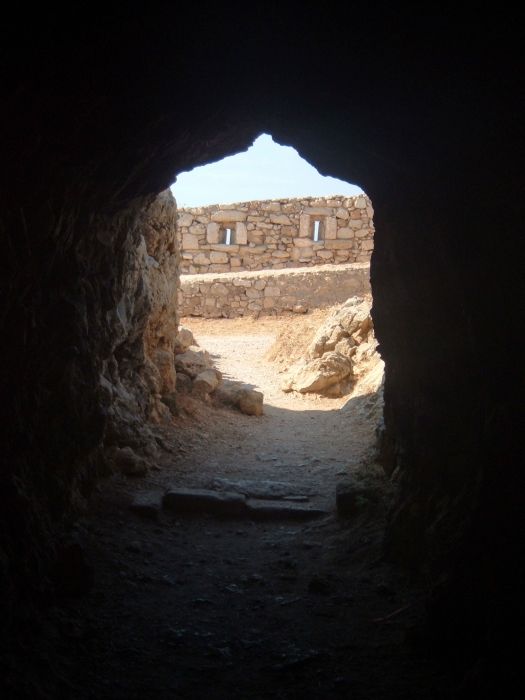 View of a doorway from inside a dark room at the Fortezza, Rethymno showing the stone walls and lookouts in the bright daylight outside.