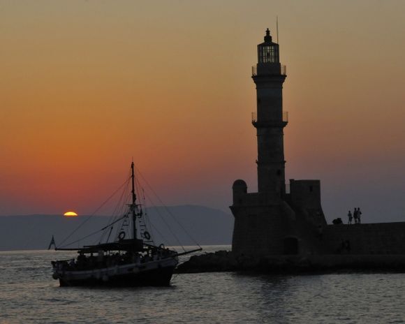 A sunset scene of Chania\'s Venetian Lighthouse and a boat entering the harbor.