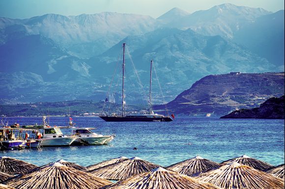 Looking south from Marathi beach across the Souda Bay harbor entrance. A large British-flagged yacht is anchored near the entrance to Loutraki Beach. Aptera castle can be seen to the right on the opposite hill overlooking the harbor entrance with the White Mountains (Lefka Ori) as a backdrop.