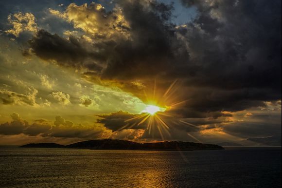 Sunrise through the clouds over Agioi Pantes island by Agios Nikolaos.