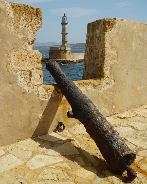 A cannon at a Firkas Fortress wall opening. Across the entrance of the harbor is the Venetian lighthouse of Chania.